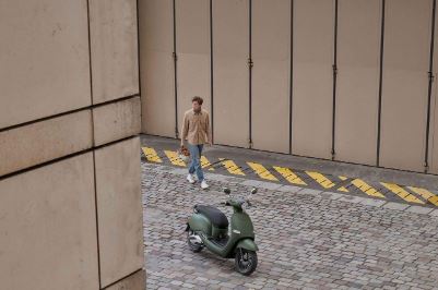 Homme avec casque à la main et s’approchant d’un scooter Brumaire vert garé sur une route pavée